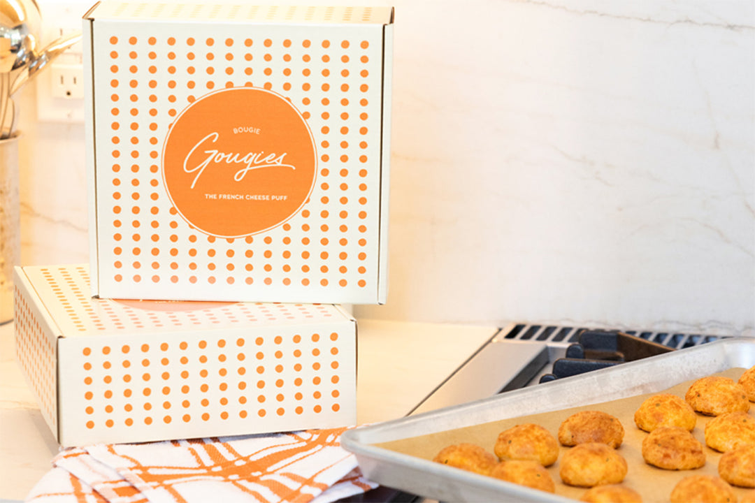 Two stacked boxes of shipped Bougie Gougies, juxtaposed with the French cheese puffs baked to golden perfection on a baking sheet